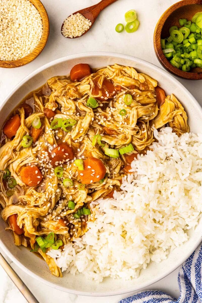 white bowl of white rice and chicken and carrots garnished with scallions and sesame seeds, surrounded by bowls of scallions and sesame seeds