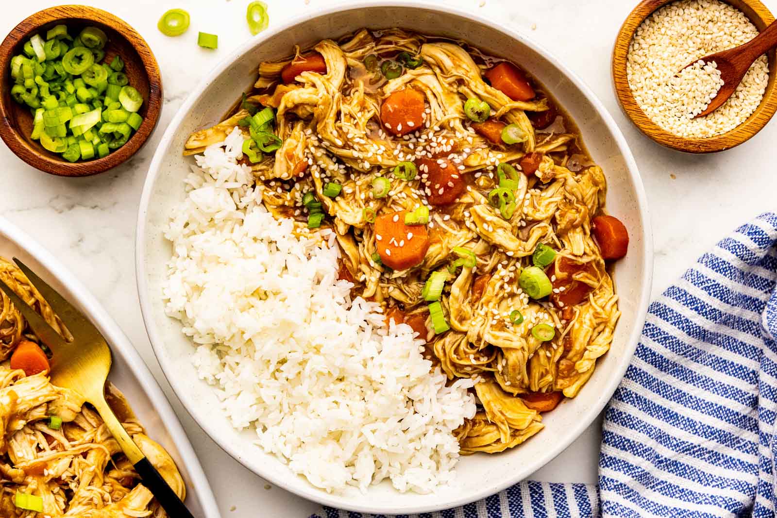 overhead view of white dinner bowl of chicken and carrots with rice surrounded by bowls of sesame seeds and scallions, and platter of chicken and carrots with fork