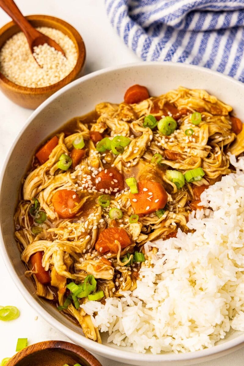 overhead view of chicken and carrots garnished with scallions and sesame seeds with rice with bowls of scallions and sesame seeds on side