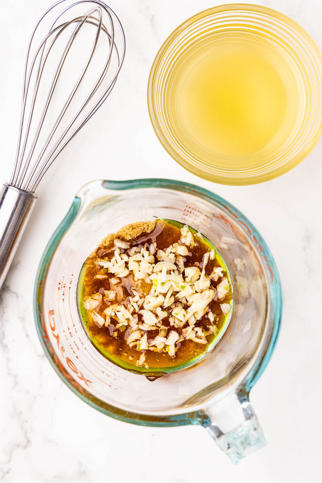 glass measuring cup with marinade ingredients and container of chicken broth with wire whisk on side