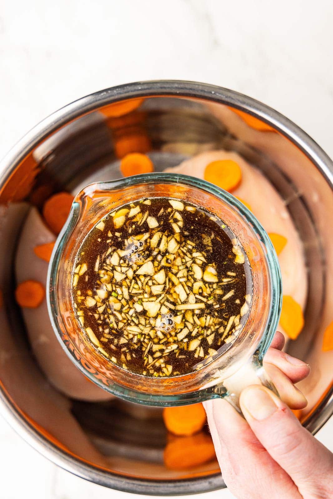 glass measuring cup of marinade being held over instant pot bowl with carrots and chicken