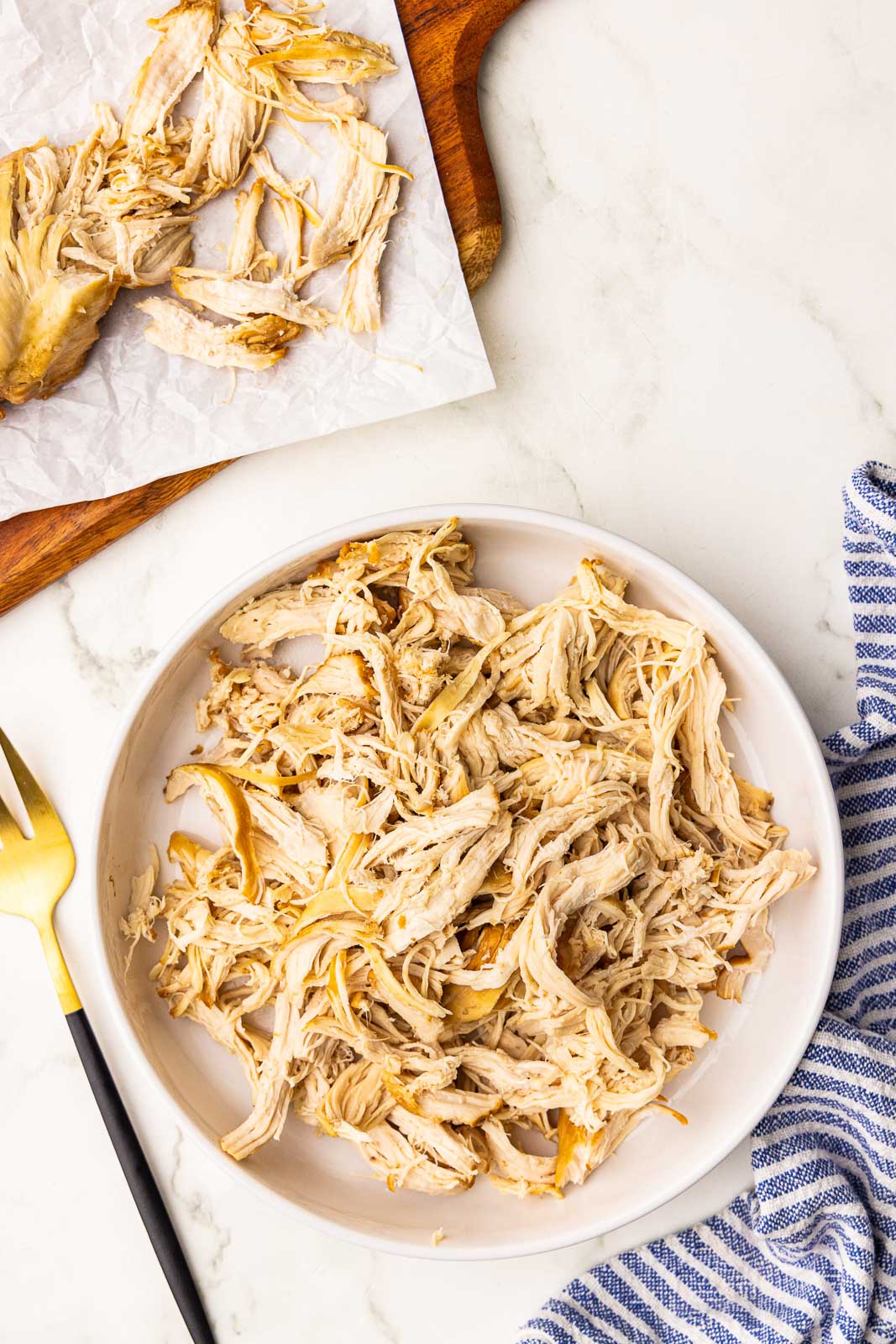 white bowl containing shredded chicken with fork on side, along with parchment lined cutting board with shredded chicken