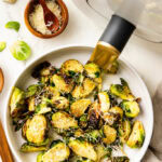overhead view of white platter of Brussels sprouts with wooden spoon, Parmesan cheese and air fryer in background