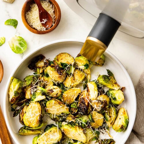 overhead view of white platter of Brussels sprouts with wooden spoon, Parmesan cheese and air fryer in background