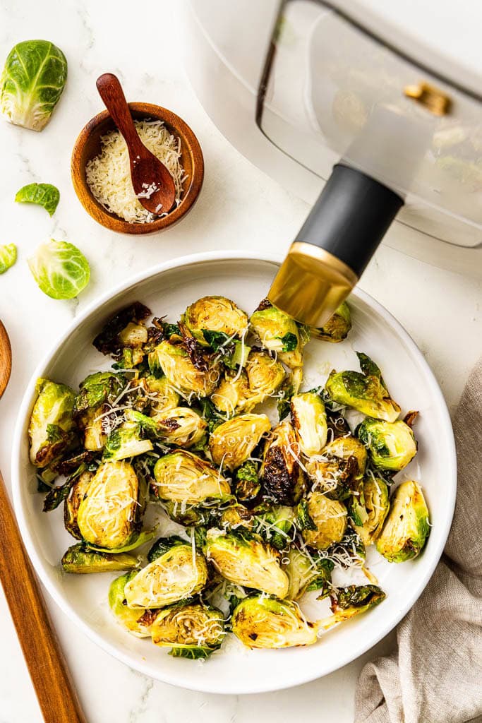 overhead view of white platter of Brussels sprouts with wooden spoon, Parmesan cheese and air fryer in background