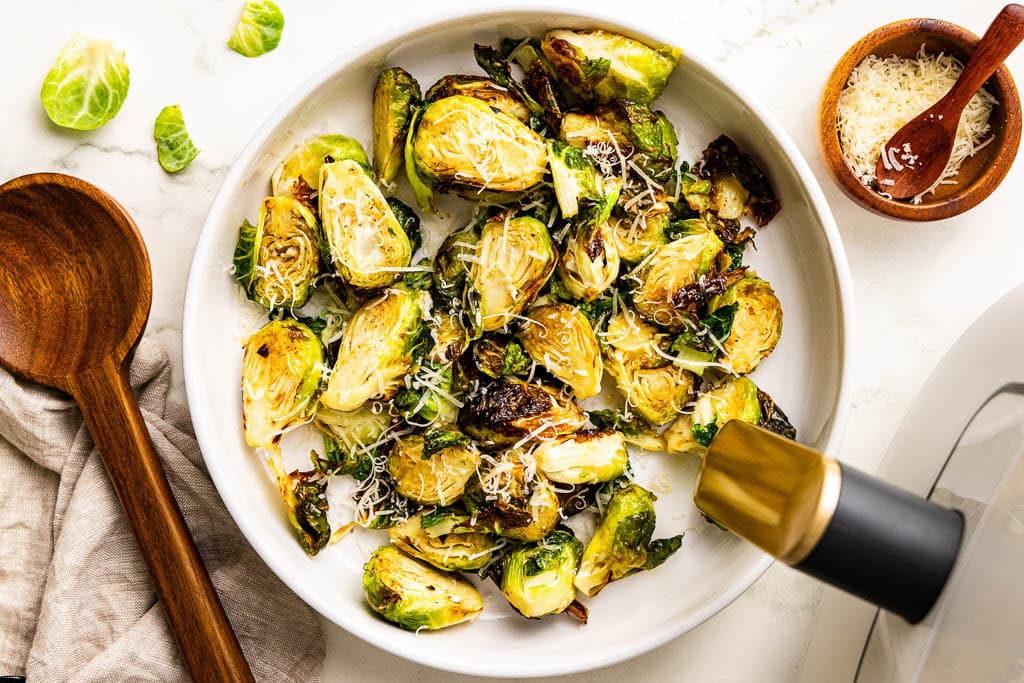 overhead view of white platter of Brussels sprouts with wooden spoon, air fryer and container of Parmesan cheese on side