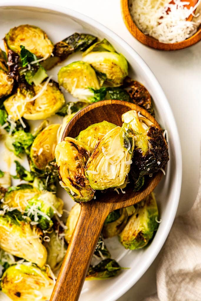 closeup of wooden spoon of Brussels sprouts with white platter of Brussels sprouts and container of Parmesan cheese in background