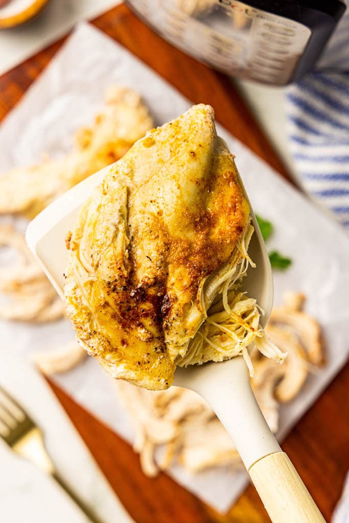 closeup of spatula holding cooked chicken with parchment lined wooden cutting board with chicken and instant pot in background