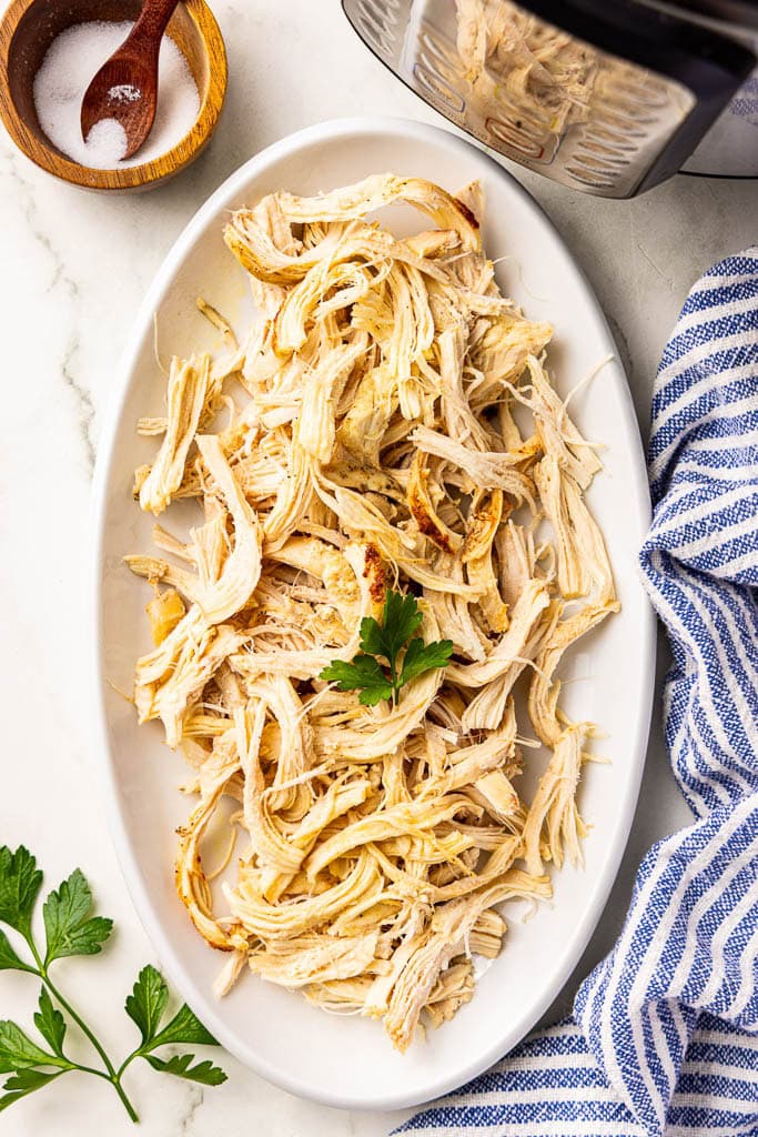 overhead view of shredded chicken breast on white platter with salt container and instant pot in background