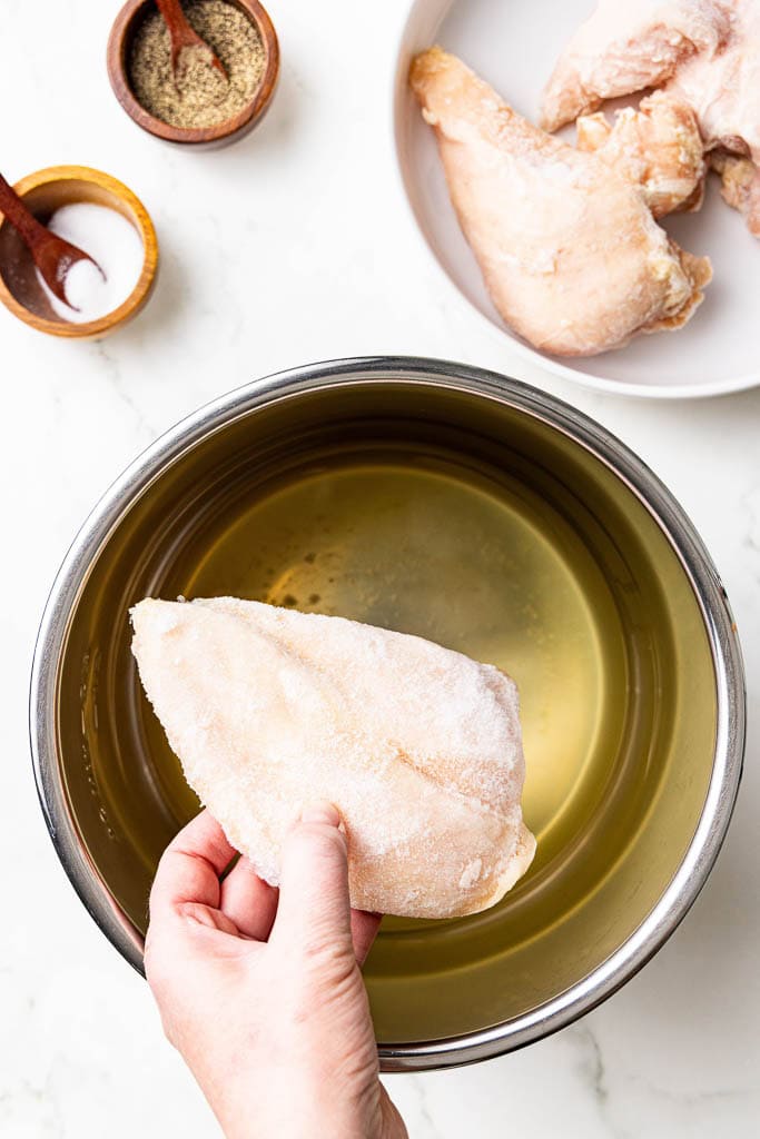 chicken breast being added to instant pot with chicken broth with bowl of chicken breasts and salt and pepper on side