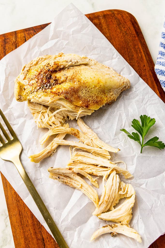 parchment lined wooden cutting board with chicken breast being shredded with fork