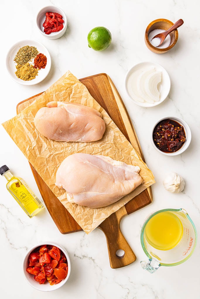 ingredients to include parchment lined wooden cutting board with 2 chicken breasts, dry seasonings, tomato paste, whole lime, salt, yellow onion, chipotle peppers in adobo sauce, garlic cloves, chicken broth, fire roasted diced tomatoes, and olive oil.