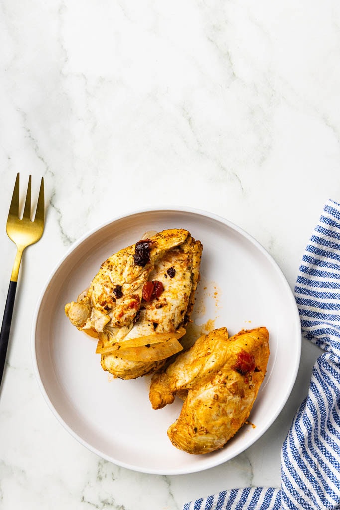 white plate with 2 cooked chicken breasts with gold fork and tea towel on side