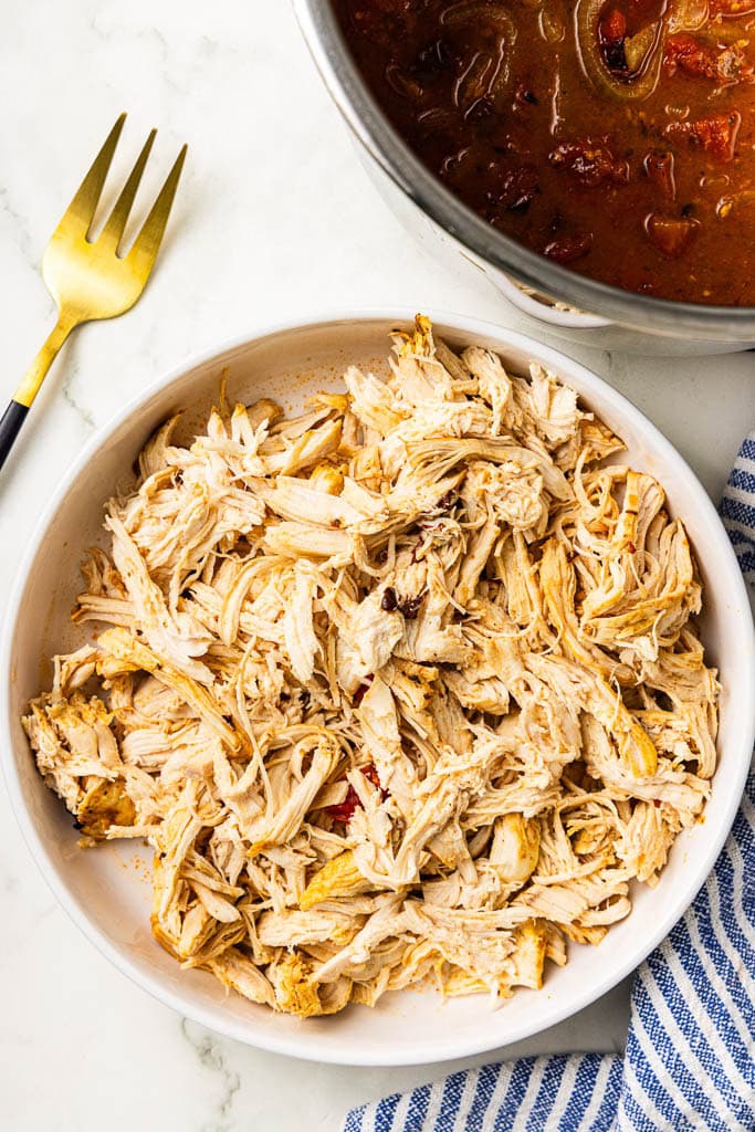 white plate with shredded chicken breasts with instant pot with veggies and gold fork on side
