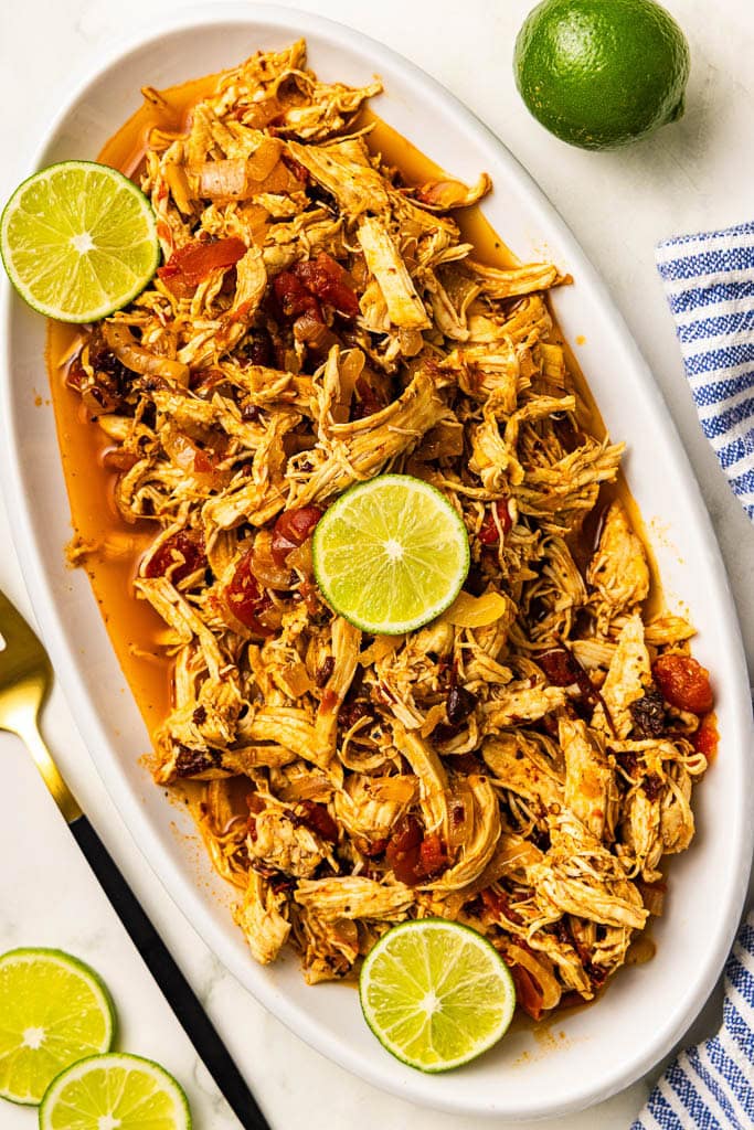 long white platter of chicken tinga with 3 lime slices on top, with lime and lime slices and gold fork on side