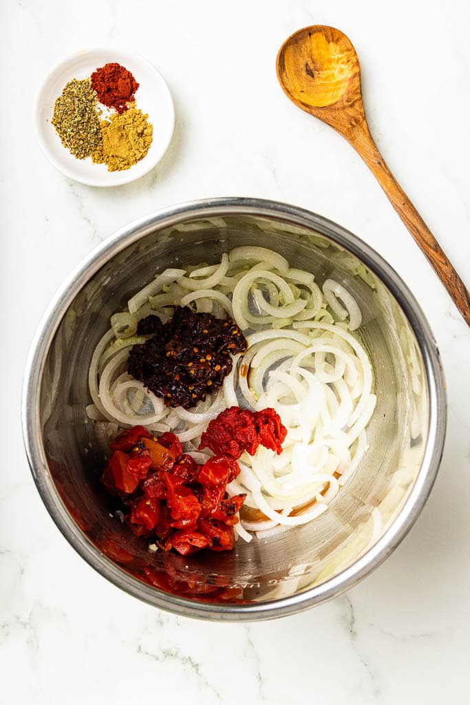 overhead view of instant pot with onion, tomato paste, fire roasted tomatoes and tomatoes with seasonings and wooden spoon on side