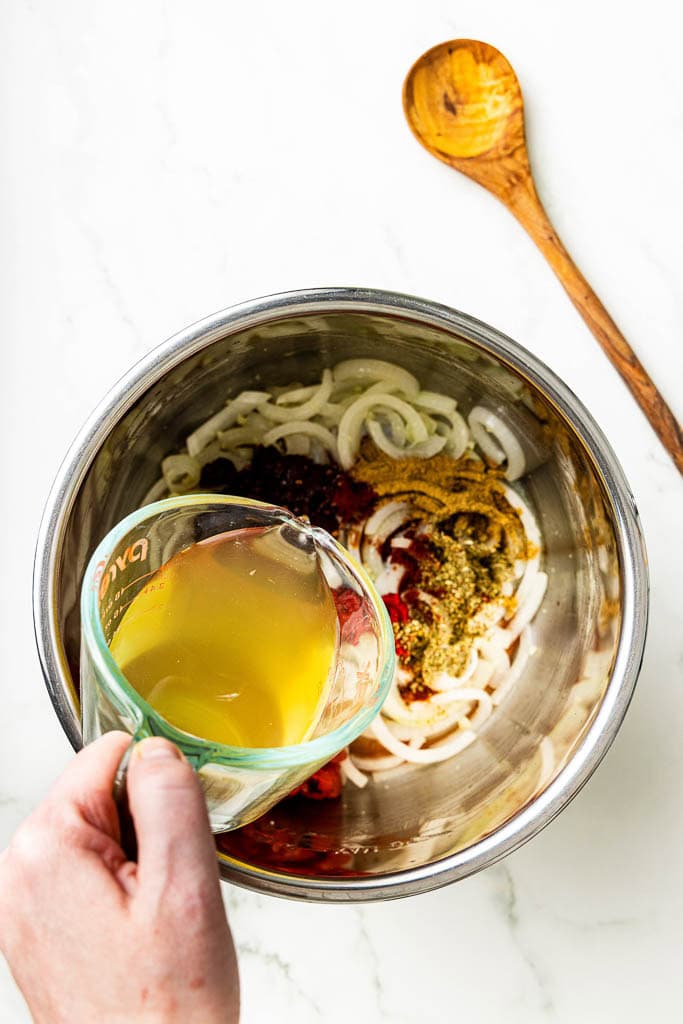 overhead view of instant pot with onion, tomato paste, fire roasted tomatoes and tomatoes, seasonings with hand pouring chicken broth into pot, with wooden spoon on side