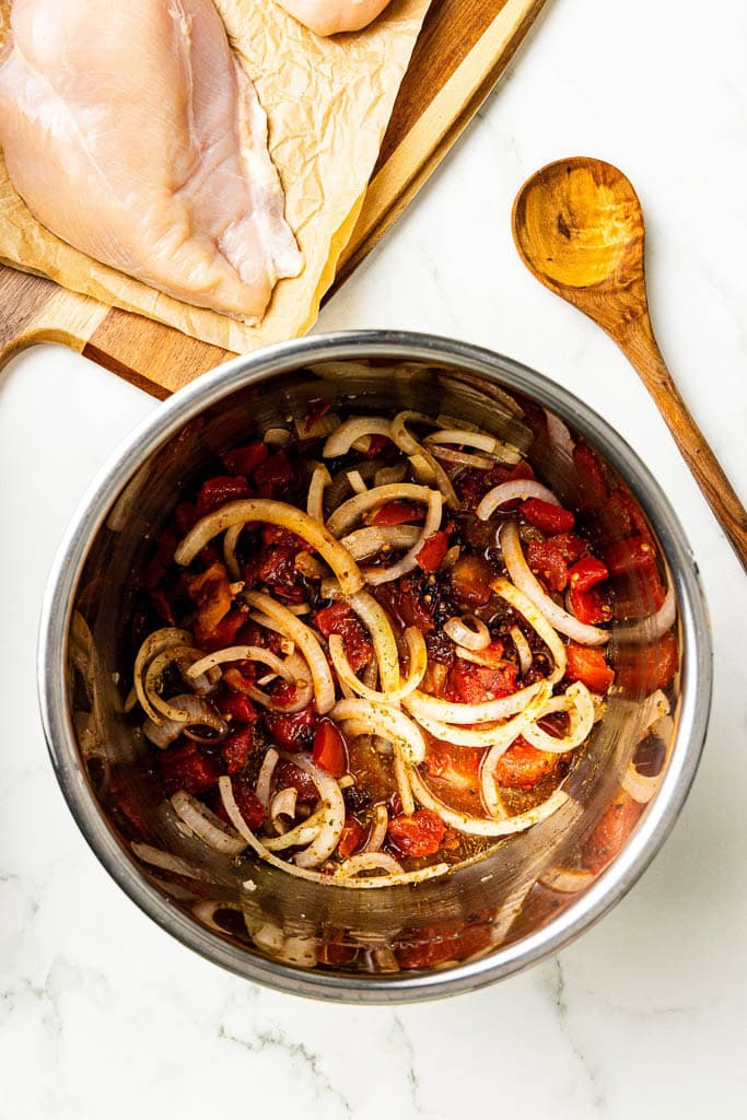 all vegetable ingredients in instant pot with wooden cutting board with chicken breasts and wooden spoon on side