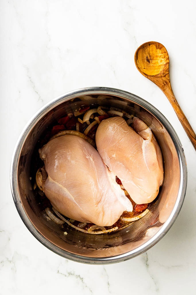 instant pot with all ingredients with 2 chicken breasts on top, with wooden spoon on side