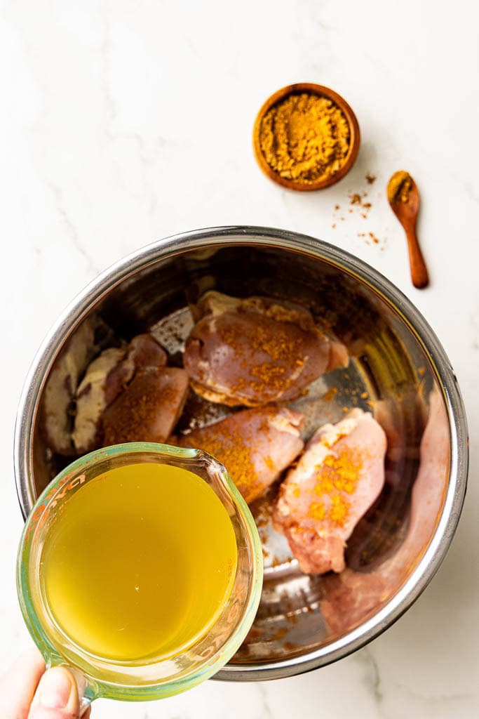 hand holding chicken broth being poured into instant pot containing seasoned chicken thighs with taco seasoning on side