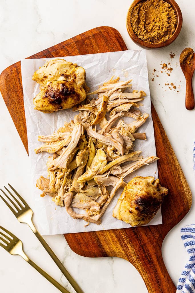parchment lined wooden cutting board with chicken thighs being shredded with 2 forks and taco seasoning on side