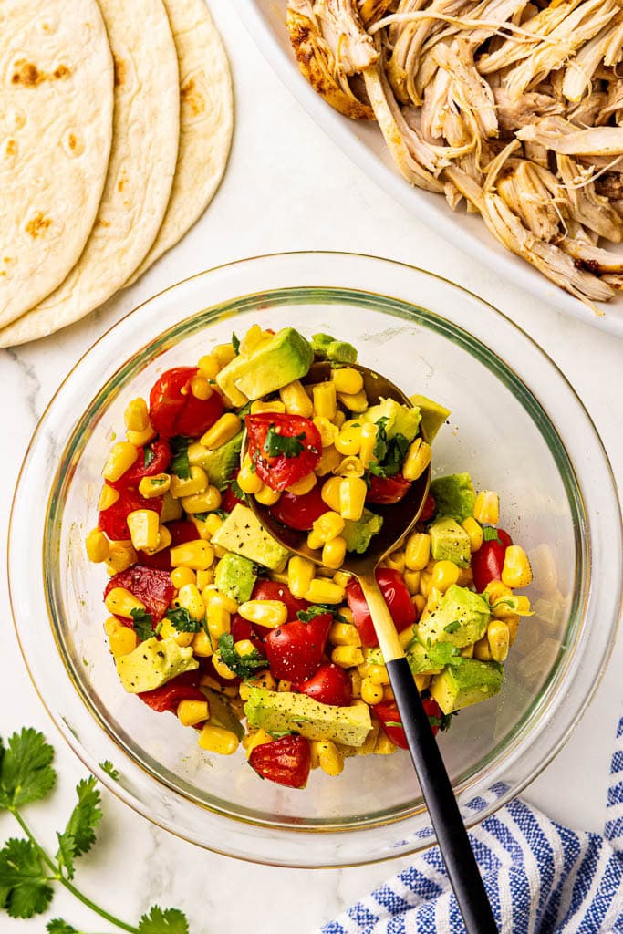 glass bowl of salsa with spoon with shredded chicken and tortillas in background