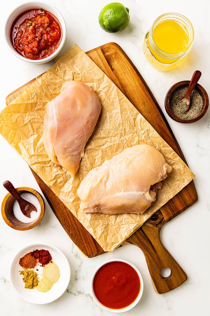 ingredients to include parchment lined wooden cutting board with 2 chicken breasts surrounded by containers of salsa, lime, chicken broth, pepper, tomato sauce, seasonings and salt