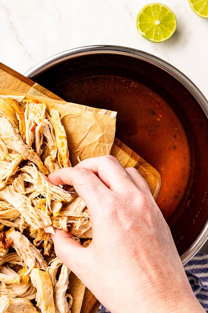 handing holding cutting board with shredded chicken being put in instant pot with tomato mixture
