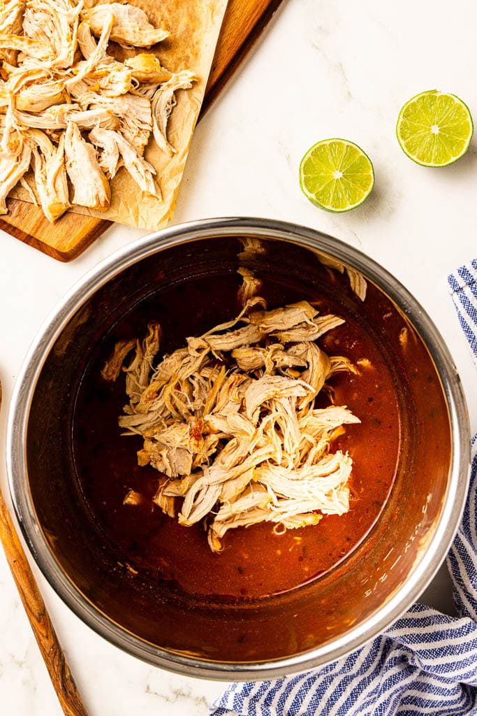 overhead view of instant pot with chicken in tomato mixture with wooden cutting board of shredded chicken and lime halves in background, and wooden spoon on side