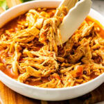 closeup of white bowl of Mexican chicken with tongs on wooden cutting board with limes in background