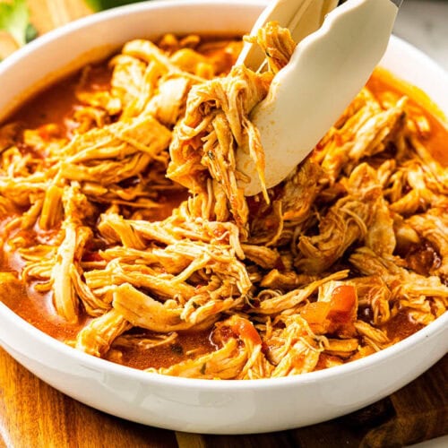 closeup of white bowl of Mexican chicken with tongs on wooden cutting board with limes in background