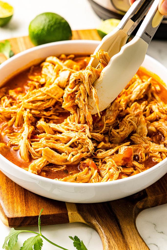 closeup of white bowl of Mexican chicken with tongs on wooden cutting board with limes in background