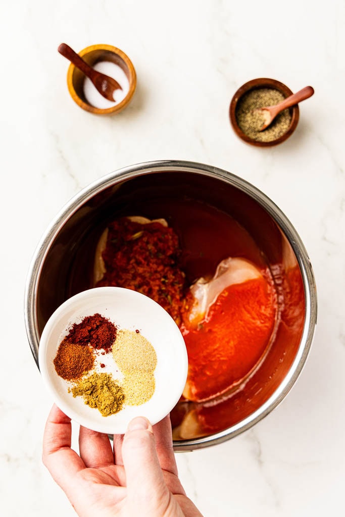hand holding plate of seasonings over instant pot with chicken, salsa and tomato sauce, with salt and pepper on side