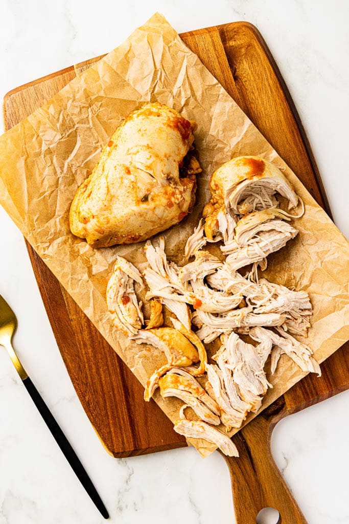 parchment lined wooden cutting board with one whole and one shredded chicken breast with fork on side
