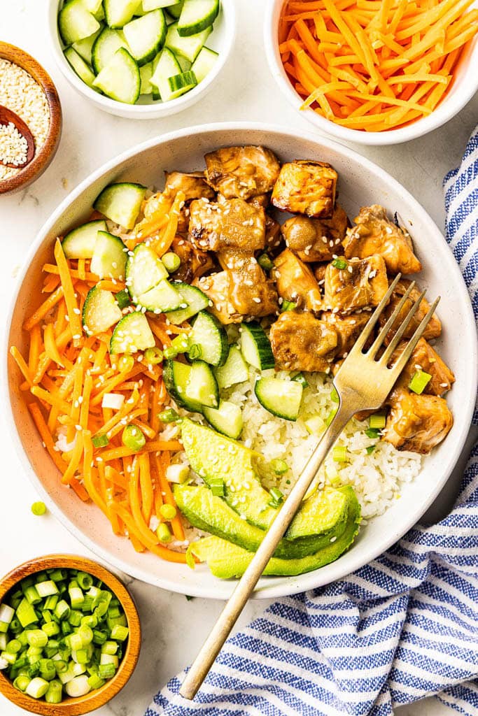 overhead view of dinner plate with salmon bites, rice, cucumber, carrots, avocado and sesame seeds, with a dinner fork, with containers of sesame seeds, cucumbers, carrots, and green onion on the side