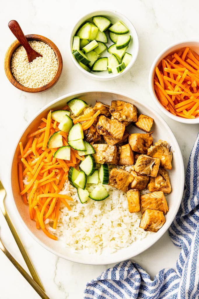 overhead view of white dinner plate of air fried salmon bites, rice, cucumber and carrots, with containers of sesame seeds, diced cucumbers and shredded carrots, with 2 forks on the side