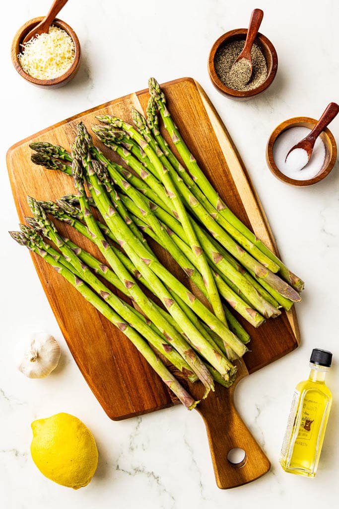 ingredients to include asparagus on wooden cutting board surrounded by containers of Parmesan cheese, black pepper, salt, olive oil, lemon and garlic
