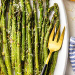 closeup view of white serving platter with baked asparagus sprinkled with Parmesan cheese and 2 lemon slices, surrounded by container of Parmesan cheese, and spoon, dish towel, and serving fork