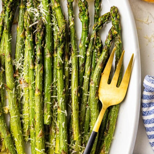 closeup view of white serving platter with baked asparagus sprinkled with Parmesan cheese and 2 lemon slices, surrounded by container of Parmesan cheese, and spoon, dish towel, and serving fork
