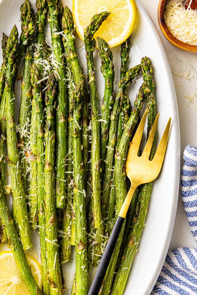 closeup view of white serving platter with baked asparagus sprinkled with Parmesan cheese and 2 lemon slices, surrounded by container of Parmesan cheese, and spoon, dish towel, and serving fork