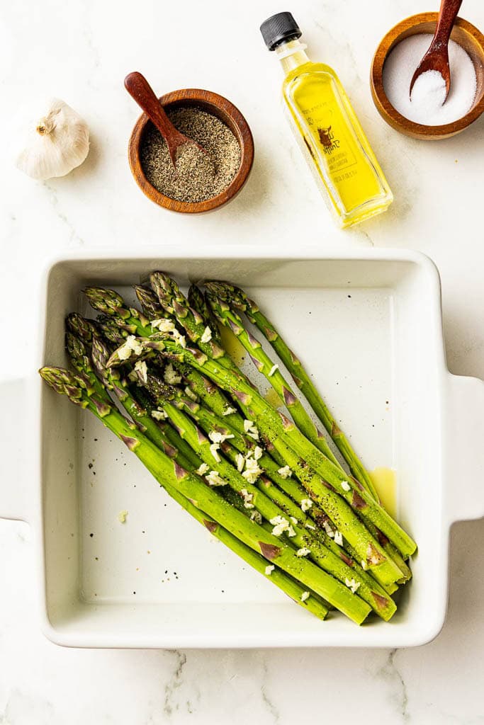 white dish containing asparagus sprinkled with minced garlic, salt, pepper and drizzled with olive oil