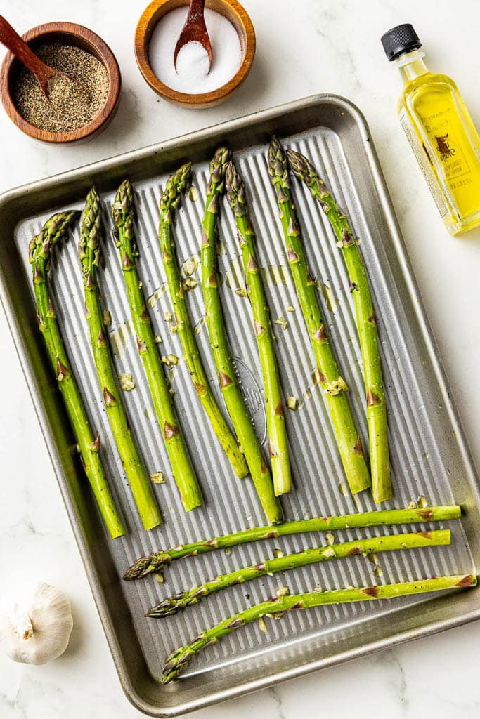 Baking sheet with asparagus surrounded by containers of salt and pepper, olive oil and garlic clove