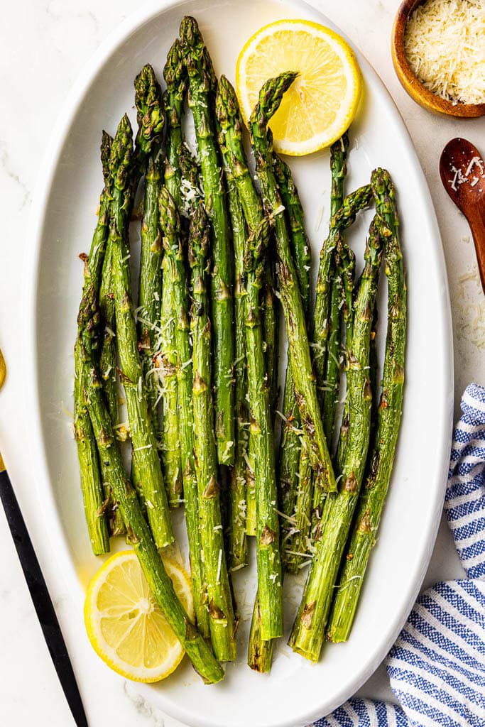 white serving platter with baked asparagus sprinkled with Parmesan cheese and 2 lemon slices, surrounded by container of Parmesan cheese, dish towel, a wooden spoon and serving fork