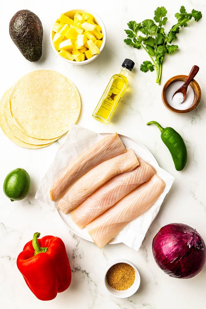 ingredients to include 4 mahi mahi fillets on white plate, surrounded by bottle of olive oil, containers of Cajun seasoning, a lime, white bowl of diced pineapple, bell pepper, red onion, jalapeno, cilantro, salt, tortillas, and avocado