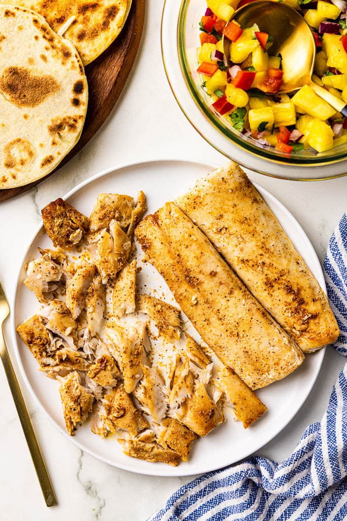 white dinner plate with mahi mahi, half broken into chunks, wooden cutting board with charred tortillas, and glass bowl of salsa on side