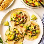 white dinner plate with filled tortillas with 2 lime wedges, bowl of salsa with spoon, and plate with mahi mahi fillets