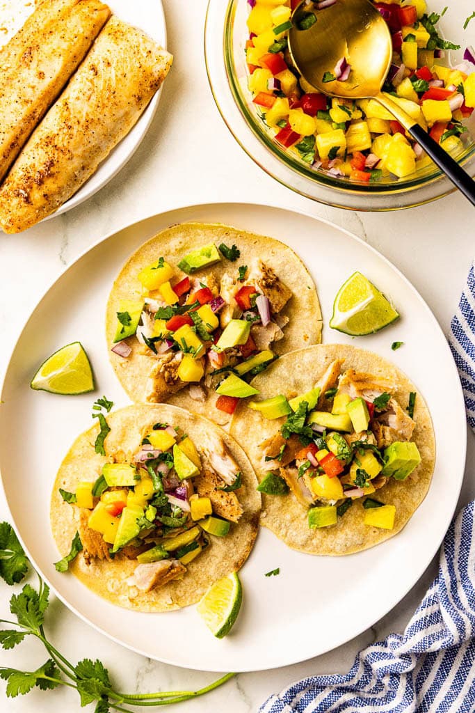 white dinner plate with filled tortillas with 2 lime wedges, bowl of salsa with spoon, and plate with mahi mahi fillets
