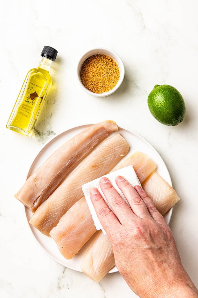 white plate with 4 mahi mahi fillets being dried with a paper towel surrounded by bottle of olive oil, Cajun seasoning and a lime