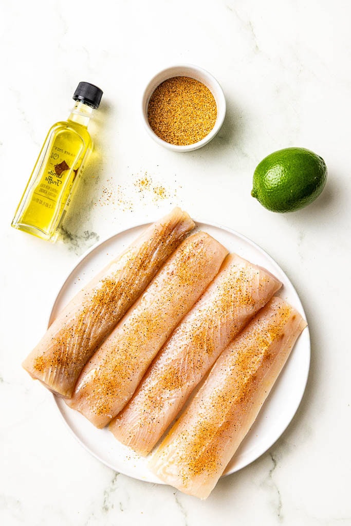 white dinner plate with 4 prepped mahi mahi fillets, surrounded by a bottle of olive oil, Cajun seasoning and a lime