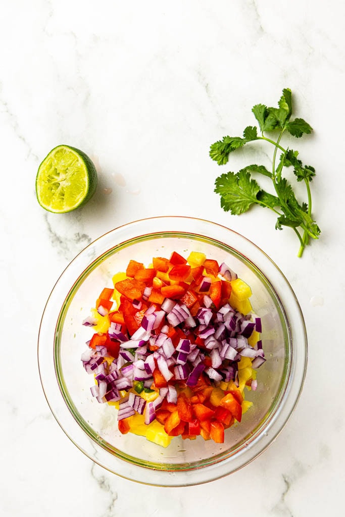 glass bowl with all fresh veggies and fruit surrounded by half of a lime and cilantro